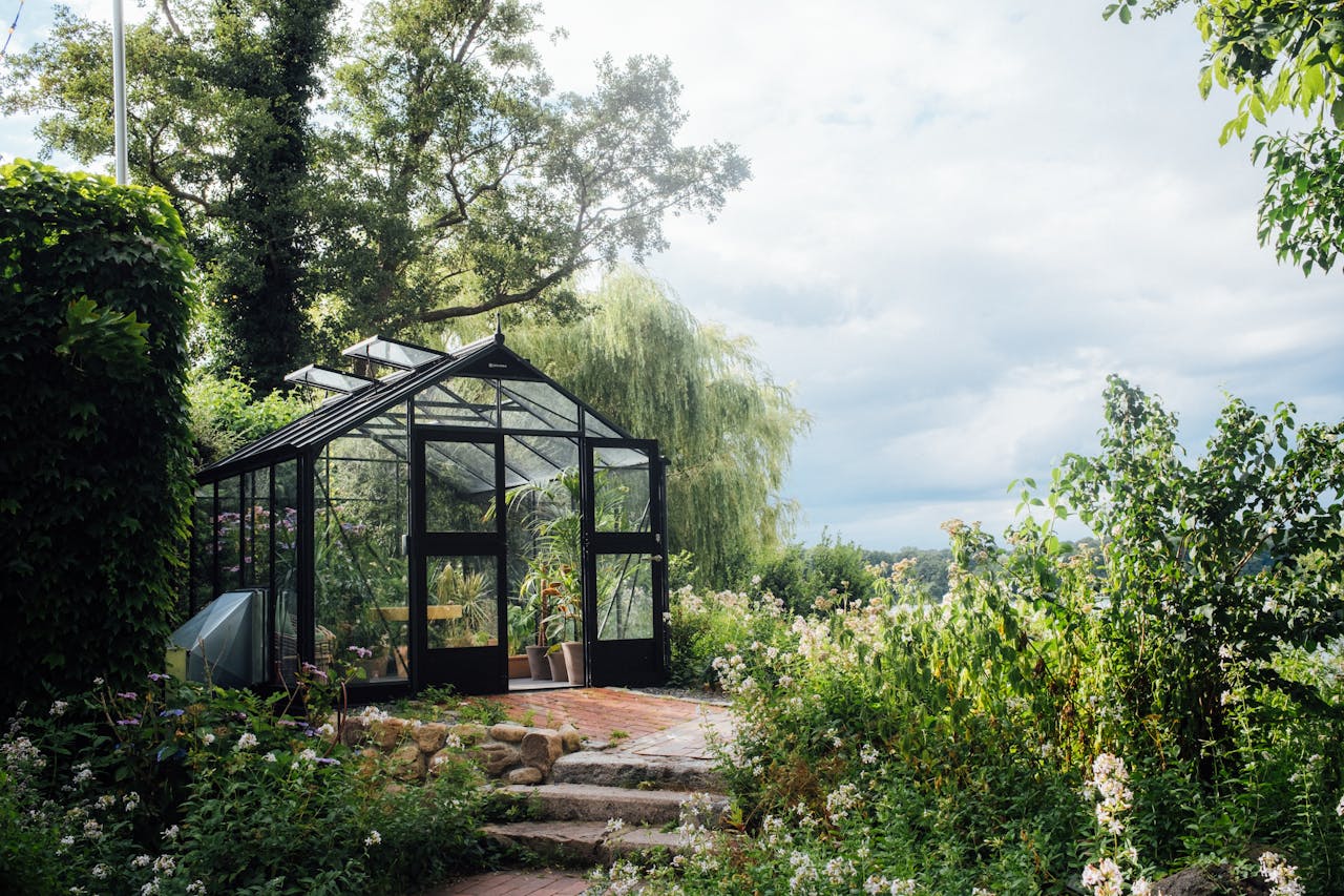 About A picturesque greenhouse nestled in a vibrant garden surrounded by greenery and trees under a bright sky.